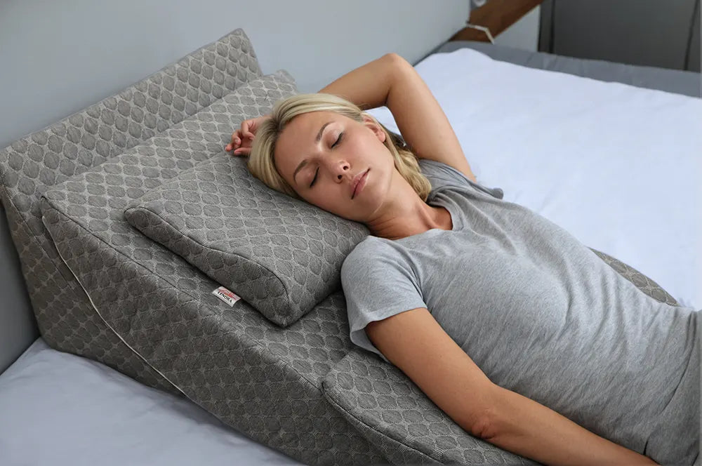 Blonde woman resting on a grey wedge pillow, eyes closed, wearing a grey t-shirt, relaxing in bed.