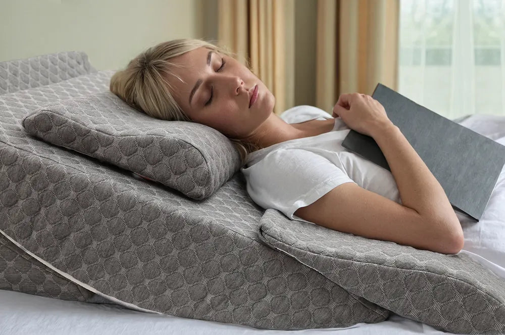 Blonde woman resting on a grey wedge pillow, holding a book on her chest, eyes closed, relaxing in bed.