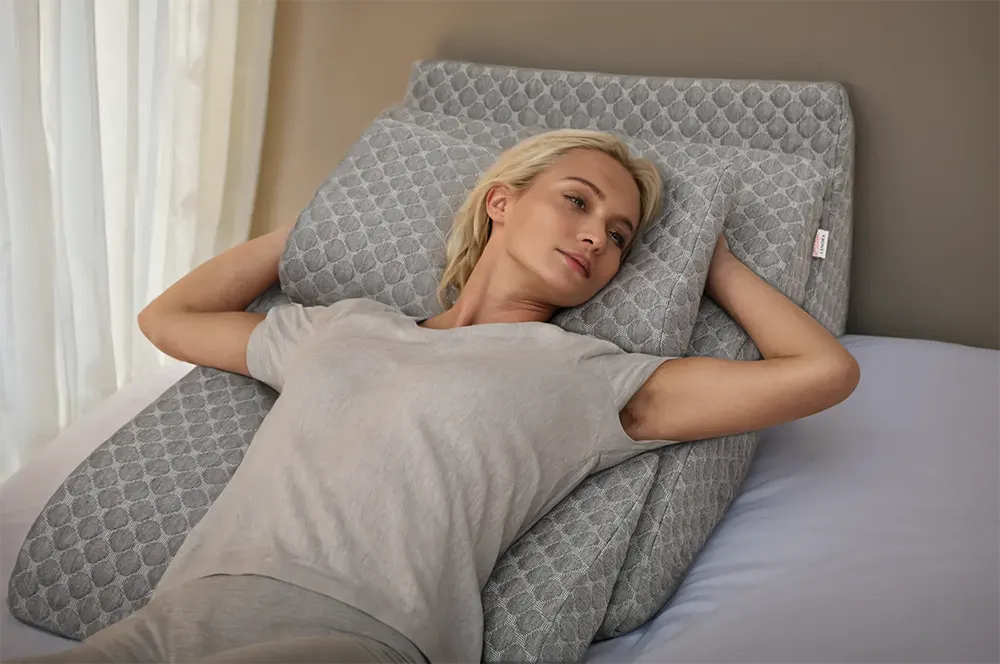 Blonde woman lying on a grey wedge pillow, arms behind her head, wearing a grey t-shirt, looking relaxed.
