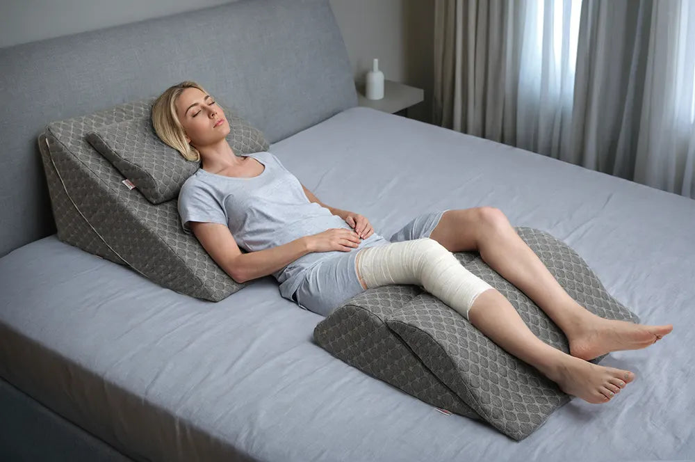 Blonde woman resting on a grey wedge pillow set, wearing a knee bandage, lying comfortably on a grey bed.
